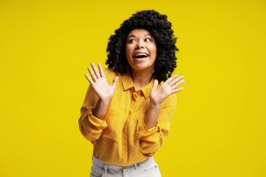 Kıvırcık saçlı, sarı gömlekli, elini kaldıran ve sarı arkaplana bakan neşeli genç kadın.