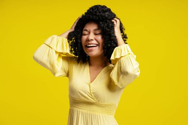 Stüdyo portresi, kıvırcık saçlı, başını tutup, gözleri kapalı gülen, neşeli, sarı bir arka planda mutluluk ve neşe ifade eden neşeli bir genç kadın.