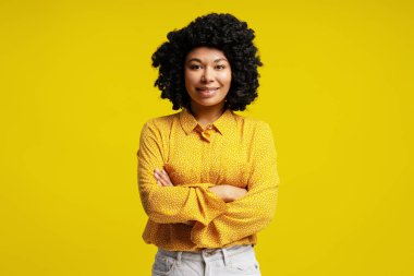 Kollarını kavuşturup gülümseyen iş kadınının stüdyo portresi, canlı sarı arka planda güven ve profesyonellik gösteriyor.