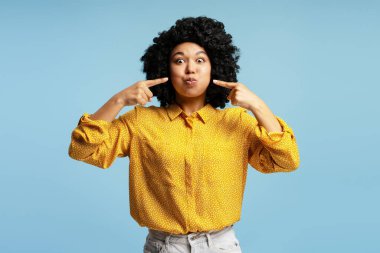 Yanaklarını gösteren kıvırcık saçlı ve mavi arka planda balık suratı yapan komik genç kadının stüdyo portresi.