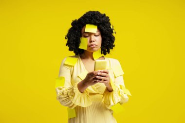 Sarı notlarla kaplı genç bir kadın sarı arka planda akıllı telefon, çoklu görev anlayışı ve hatırlatıcılar kullanıyor.