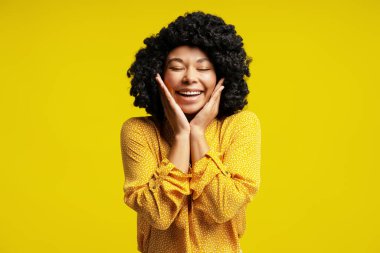 Kıvırcık saçlı neşeli bir genç kadının stüdyo portresi, sarı giyinmiş, elleriyle yüzünü tutuyor ve geniş bir gülümseme yayıyor, canlı sarı arka planda mutluluk ve heyecan yayıyor.
