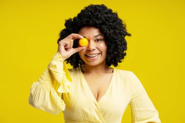 Gözünün önünde küçük sarı bir top tutan ve sarı arka planda gülümseyen neşeli Afrikalı Amerikalı kadının stüdyo portresi.