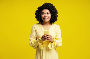 Kıvırcık saçlı, cep telefonu kullanan ve gülümseyen, sarı arka plana bakan genç bir kadının stüdyo portresi.