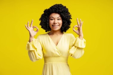 Neşeli genç bir kadının portresi. Tamam. İki elinle imzala ve parlak sarı arka planda gülümse.