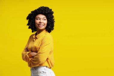 Kollarını çapraz yapmış neşeli genç bir kadının parlak sarı bir arka planda gülümseyişinin stüdyo çekimi.