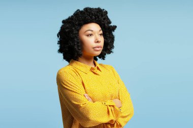 Kollarını kavuşturmuş, sarı gömlekli, mavi bir arka plana bakan kendine güvenen genç bir kadının stüdyo portresi.