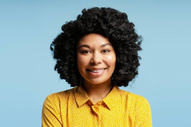 Sarı tişörtlü, mavi arka planda poz veren, kıvırcık saçlı neşeli genç bir kadının stüdyo portresi.