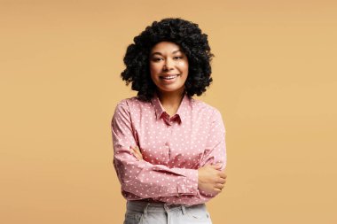 Kollarını çapraz bağlamış pembe gömlekli, bej arka planda poz veren neşeli Afro-Amerikan kadının stüdyo portresi.