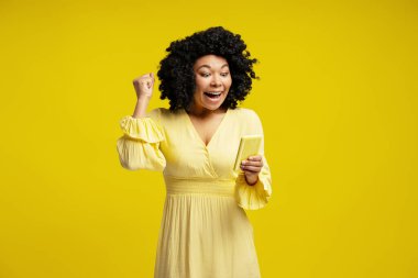 Sarı elbiseli heyecanlı Afrikalı Amerikalı kadın sarı arka planda cep telefonu kullanırken başarıyı kutluyor.