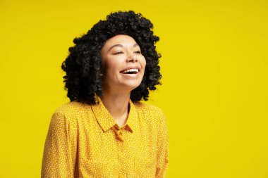 Kıvırcık saçlı neşeli bir genç kadının canlı sarı bir arka planda izole bir şekilde gülüp baktığı bir resim.