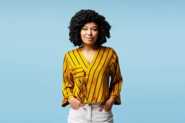 Kıvırcık saçlı ve sarı çizgili tişörtlü genç bir kadının stüdyo portresi, elleri rahat bir şekilde cebine yerleştirilmiş, canlı mavi bir arka plana karşı.