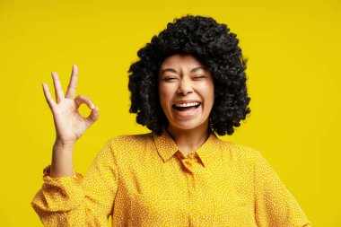 Kıvırcık saçlı neşeli genç bir kadın parmaklarıyla işaret veriyor ve sarı arka planda gülüyor.