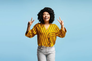 İki eliyle barış işareti gösteren, gülümseyen ve mavi arka planda günlük kıyafetler giyen neşeli genç bir kadının stüdyo portresi.