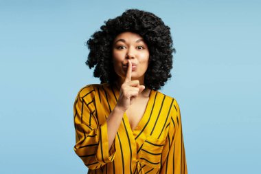Siyah kıvırcık saçlı ve sarı çizgili gömlekli genç bir kadının stüdyo portresi açık mavi arka planda parmaklarıyla sessiz bir jest yapıyor.