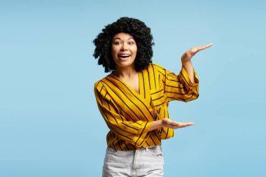 Heyecanlı, kıvırcık saçlı, elleri arasında ürün reklamı için yer gösteren, mavi arka planda izole edilmiş Afrikalı Amerikalı kadın.