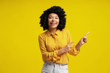 Afro-Amerikalı neşeli bir kadının işaret parmaklarıyla yanlamasına gösteren bir stüdyo portresi, sarı arka planda bir şeylerin reklamını yapıyor.