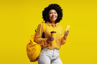 Neşeli genç öğrenci cep telefonu kullanıyor ve kahve içiyor, sarı gömlek ve sırt çantası giyiyor, sarı arka planda duruyor.