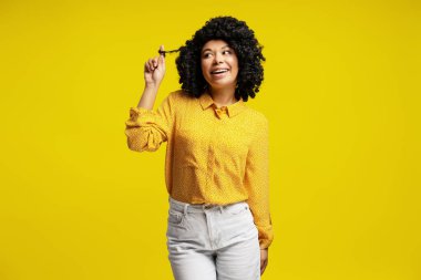 Saçlarıyla oynayan ve sarı bir arka planda gülümseyen genç bir kadının stüdyo fotoğrafı.