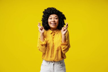 Umut dolu parmakları çapraz genç bir kadının stüdyo portresi. Günlük kıyafetler giymiş, parlak sarı bir arka planda izole edilmiş.