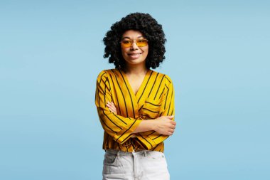 Sarı bluz ve güneş gözlüğü takmış gülümseyen Afro-Amerikan iş kadınının stüdyo portresi, mavi arka planda çapraz kollarla poz veriyor.