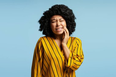 Kıvırcık saçlı, sarı çizgili gömlekli açık mavi arka planda diş ağrısı olan genç bir kadının stüdyo portresi.