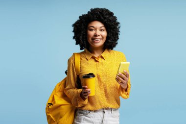 Elinde kahve ile gülümseyen ve cep telefonu kullanan genç bir kadın, sırt çantasının yanında canlı mavi bir arka plana yaslanmış.