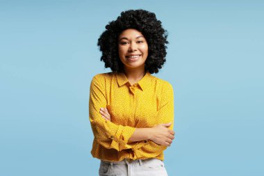 Mavi arka planda izole edilmiş, çapraz kollu sarı gömlek giyen Afro-Amerikan iş kadınının stüdyo portresi.