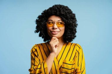 Düşünceli, kıvırcık saçlı, sarı gözlüklü ve çizgili tişörtlü Afro-Amerikan kadın çenesini tutup mavi arka plana bakıyor.