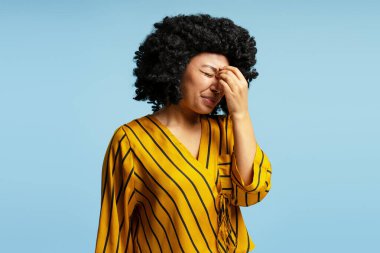 Stüdyo portresi kıvırcık saçlı, sarı çizgili gömlekli, güçlü bir baş ağrısından muzdarip bir genç kadın, açık mavi arka planda elleriyle yüzüne dokunuyor.
