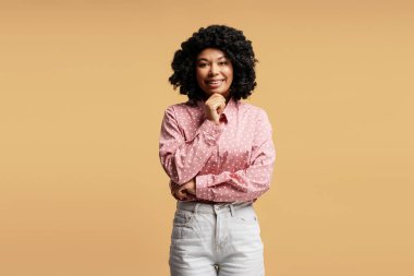 Stüdyo portresi kıvırcık saçlı, pembe noktalı gömlek ve beyaz kot pantolon giyen, gülümseyerek çenesine dokunan neşeli bir genç kadın.