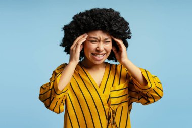 Stüdyo portresi kıvırcık saçlı ve sarı gömlekli güçlü bir baş ağrısından muzdarip, parmaklarıyla şakaklarına dokunan, sağlıksız hisseden genç bir kadın.