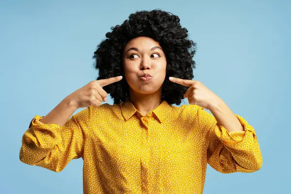 Mavi arka planda tombul yanaklarını işaret eden genç bir kadının stüdyo portresi.