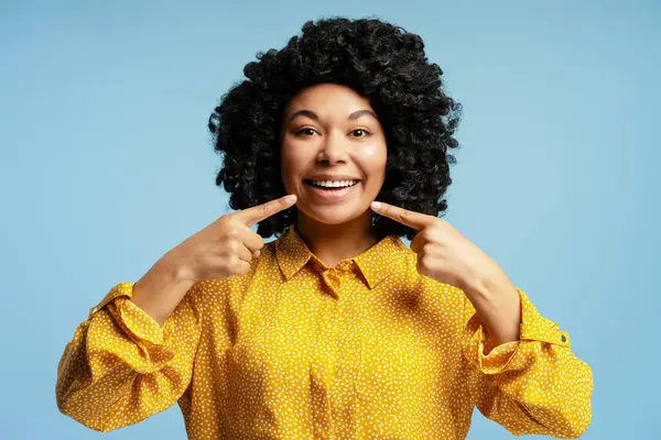 Mavi bir arka planda dururken mükemmel gülümsemesini gösteren neşeli bir kadının stüdyo portresi.