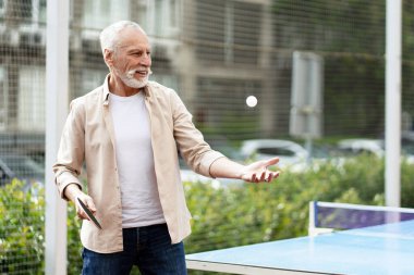 Yaşlı adam parkta pinpon oynamaktan zevk alıyor. Gülümserken ve dışarıda aktif kalırken topu ustalıkla fırlatıyor.