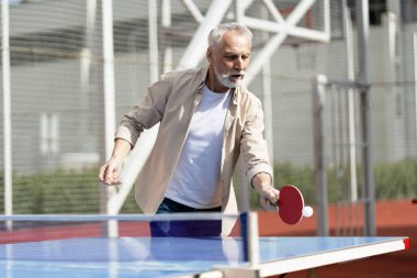 Yaşlı bir adam açık masa tenisinden hoşlanıyor, aktif bir yaşam tarzını benimsiyor ve güneşli bir yaz gününde formda kalıyor.