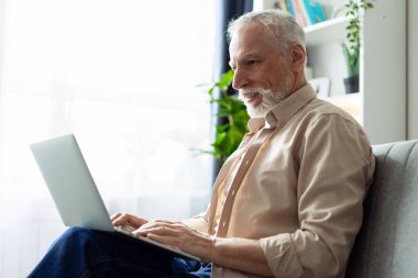 Mutlu emekli adam evdeki kanepede rahat bir şekilde oturuyor, online iletişim ve modern teknolojinin tadını çıkarmak için dizüstü bilgisayar kullanıyor.