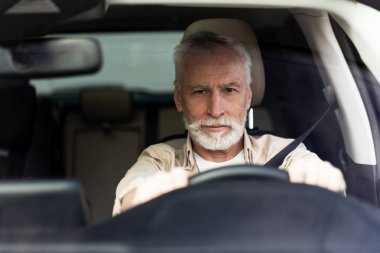 Beyaz sakallı yaşlı bir adam araba kullanıyor, günlük kıyafetler giyiyor ve bir yol gezisinde dosdoğru ileriye bakıyor.