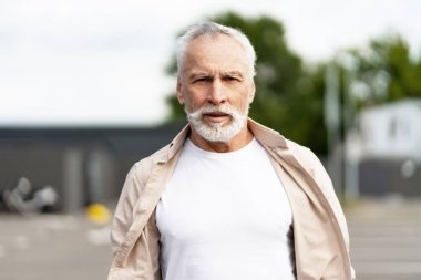 Beyaz saçlı, sakallı, günlük kıyafetler giyen, kentli bir ortamda duran, kendinden emin görünen son sınıf öğrencisi.