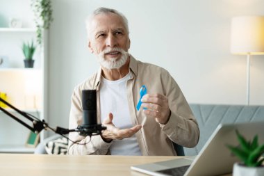 Olgun erkek etkileyicisi mavi kurdele gösteriyor, podcast 'te prostat kanserinden bahsediyor, ev stüdyosunda ses kaydı yapıyor.