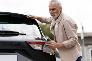 Yaşlı bir adam, aracını mikrofiber bir bezle dikkatlice temizliyor, lekesiz ve parlak bir son garantiliyor.