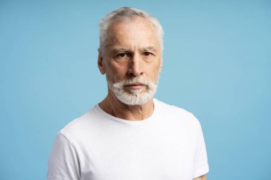 Beyaz saçlı, sakallı, beyaz tişörtlü ve mavi arka planda ciddi görünen kendinden emin olgun bir adamın portresi.