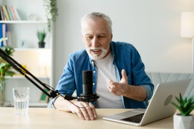 Mutlu, olgun bir adam rahat ofisinden bir podcast kaydediyor. Profesyonel bir mikrofon ve dizüstü bilgisayar kullanarak bir bardak suyun keyfini çıkarıyor.