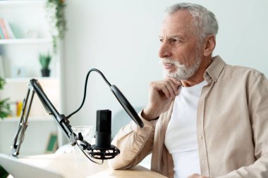 Gri saçlı ve sakallı düşünceli olgun bir adam profesyonel mikrofon kullanarak stüdyoda ses yayını yapıyor.