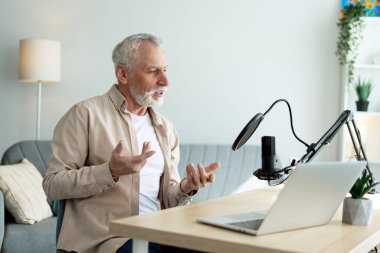 Profesyonel bir mikrofon ve dizüstü bilgisayar kullanarak rahat bir ev stüdyosunda podcast kaydeden gri saçlı bir adam.