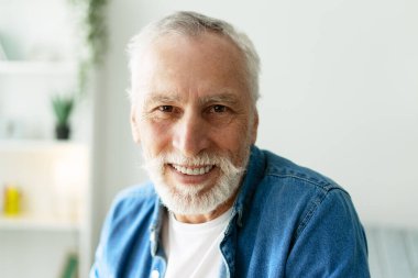 Beyaz sakallı, bıyıklı, beyaz tişörtlü, kameraya bakıp gülümseyen, bulanık bir ev arka planında gülümseyen neşeli bir son sınıf öğrencisinin portresini yakından çek.