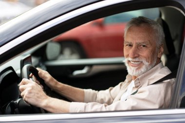 Gülümseyen kıdemli bir adam günlük kıyafetlerle araba sürüyor, şehir sokaklarında gezinirken açık yolların özgürlüğünün tadını çıkarıyor.