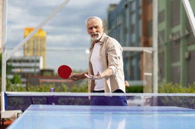 Gri saçlı adam açık havada masa tenisi oynamaktan zevk alıyor, yaz güneşinde gülümserken raketiyle topa ustaca vuruyor.