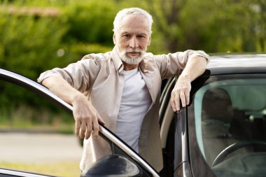 Kendinden emin kıdemli adam kapıyı açık tutarken yeni arabasına yaslanıyor, doğadaki emekliliğinin tadını çıkarıyor.