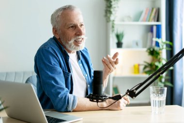 Yaşlı radyo sunucusu mikrofona konuşuyor ve evinde rahat ettiği stüdyodan podcast kaydederken hareketli bir şekilde işaret veriyor.